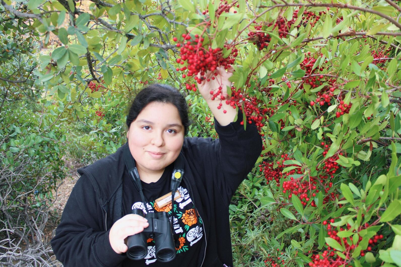 Belen surrounded by bright, red Toyon berries while holding a binocular.